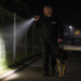 Security guard walking a dog at night using an AK-9 LED torch along a fenced pathway